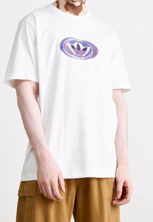Persoon draagt een wit T-shirt met korte mouwen met een vervormd blauw en paars Adidas-logo, gecombineerd met een bruine cargobroek.