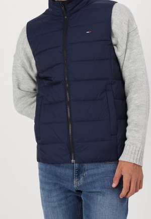 Homme portant une veste matelassée bleu marine sur un pull gris clair et un jean bleu, la veste zippée.