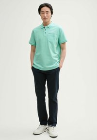 Miętowy polo z krótkim rękawem i kieszenią na piersi, w połączeniu z ciemnonawigacyjnymi spodniami i białymi trampkami. Gładka tekstura materiału.