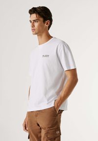 Wit katoenen T-shirt met korte mouwen, met zwarte tekstprint op de borst. Het model draagt bruine cargobroek met zakken.