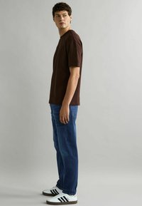 Camiseta de algodón marrón de manga corta, combinada con jeans azules. Zapatillas blancas con rayas negras a los lados. Diseño simple y casual.