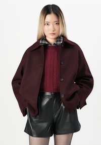 Manteau en laine bordeaux avec un grand col et des boutons noirs, superposé à un pull en laine torsadée rouge et un short en cuir noir.