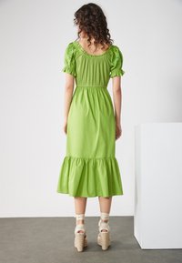 Grünes Baumwoll-Midikleid mit Puffärmeln, geraffter Taille und gestuften Rüschen am Saum. Kombiniert mit beigefarbenen, strukturierten Keil-Sandalen.
