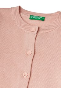 Světle růžový pletený svetr s žebrovaným límcem, který má tři kulaté knoflíky a zelenou nálepku s nápisem "United Colors of Benetton."