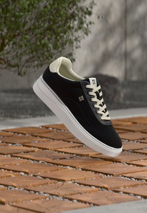 Zwart-witte veter sneakers met witte zool en "HUGO" branding, zwevend boven een patroon van houten en stenen grond buiten.