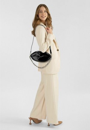 Borsa in pelle verniciata nera a forma di labbra, con manico a catena e design elegante. Indossata con un completo crema e tacchi marroni.