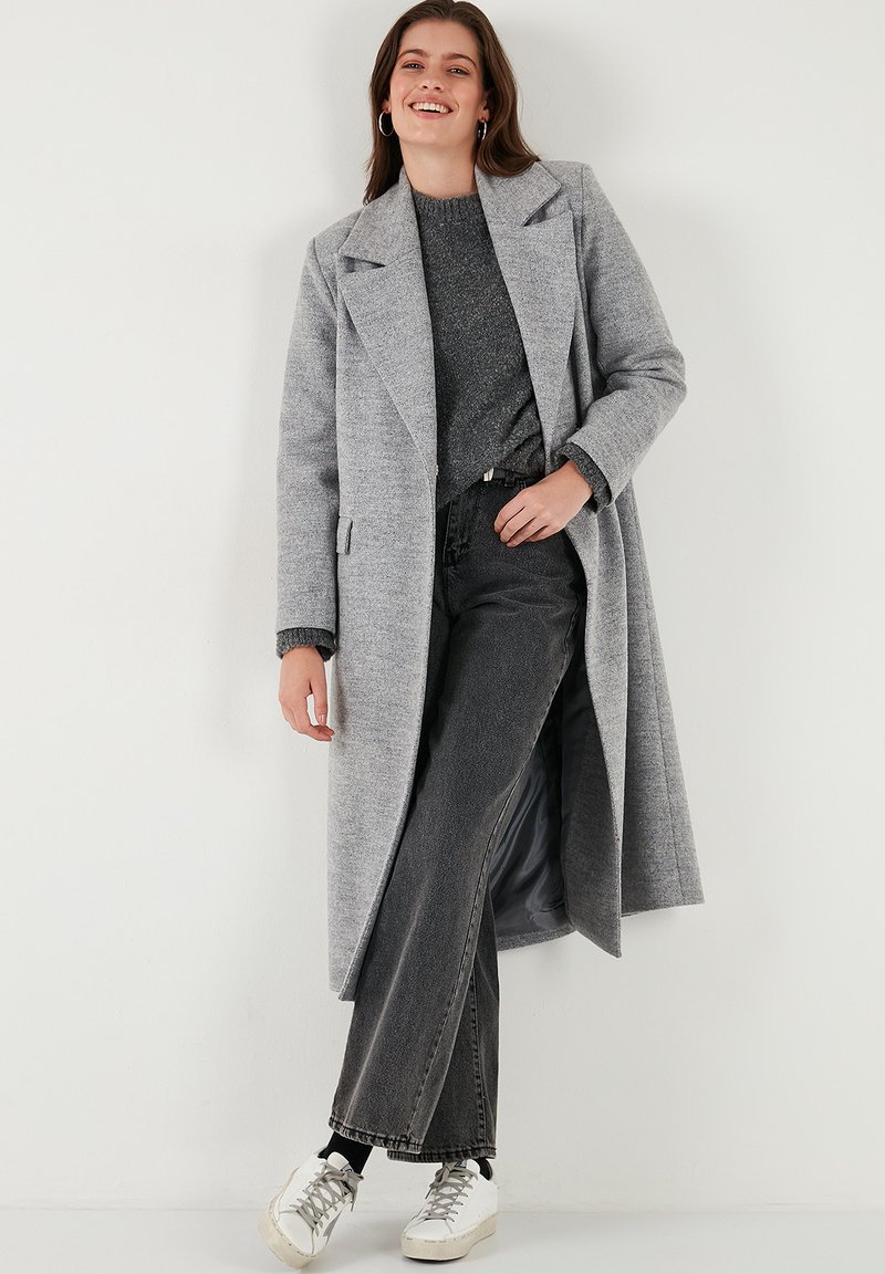 Gray long wool coat with wide lapels, worn over a gray textured sweater and black flared jeans. White sneakers complete the outfit.