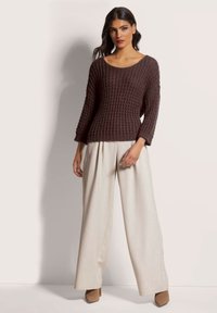 Braun strukturierter Strickpullover mit weitem Ausschnitt, kombiniert mit lockeren beigen Hosen und beigen High Heels, vor einem hellen Hintergrund.