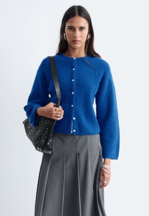 Femme portant un cardigan bleu boutonné et une jupe plissée grise, tenant un sac à main noir tressé sur l'épaule, sur un fond uni.