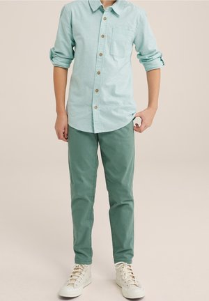 Garçon portant une chemise à boutons bleu clair, un pantalon vert et des baskets montantes blanches, debout devant un fond uni.