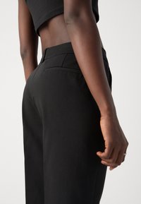 Personne portant un pantalon taille haute noir et un crop top noir, debout avec une main détendue le long du corps.