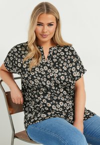 Blusa negra con flores blancas y acentos marrones. Tejido ligero, mangas cortas y escote en pico. Combinada con vaqueros azules.