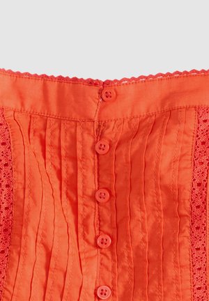Jasnopomarańczowa tkanina z pionowymi plisami, koronkową obwódką po bokach oraz pionowym rzędem sześciu pomarańczowych guzików na środku.