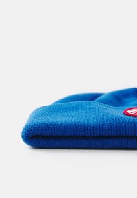 Mėlynas megztas kepurė su perlenkimu ir apvalia raudona etikete šone. Tekstūruota medžiaga, be siūlių dizainas ir minkštas išvaizda.