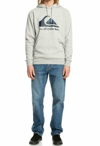 Sudadera con capucha gris con un logo impreso en negro y azul, que cuenta con capucha con cordón, bolsillo tipo canguro y puños acanalados. Combinada con vaqueros azules.