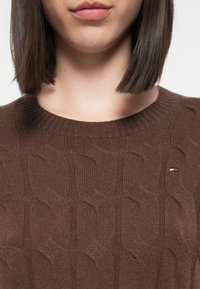Brown knitted sweater featuring a textured wavy pattern and a small logo in red, white, and blue on the chest. Round neckline.