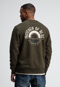 Militair groene sweatshirt met een ronde hals. Bevat een grote grafische print op de achterkant met "BUTCHER OF BLUE" in het wit. Geribbelde manchetten en zoom.