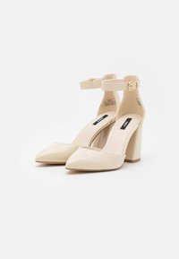 Beige kiilto nahasta valmistetut korkokengät, joissa on terävä kärki, nilkkahihna ja palikkakorko. Tyylikäs muotoilu, jossa on sileä rakenne.