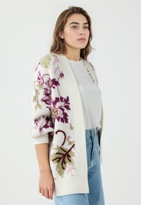 Strickjacke in Creme mit lila, grünem und pinkem Blumenstickerei. Lockere Passform, ¾-Ärmel, weiche Textur und offenes Frontdesign.
