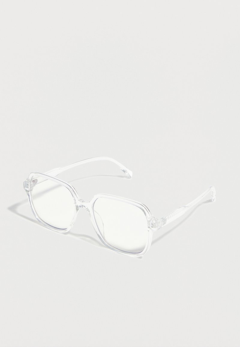 Διάφανα γυαλιά με τετράγωνο πλαίσιο και διαυγή φακούς, τοποθετημένα σε ανοιχτό γκρι επιφάνεια, με διακριτικές σκιές.