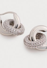 Pendientes de aro plateados con diseños circulares entrelazados. Un aro está adornado con piedras claras y brillantes, creando un acabado texturizado.