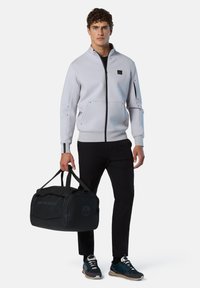 Felpa grigia chiara con zip e tasche, abbinata a pantaloni neri e sneakers, mentre tiene una borsa a tracolla nera con il logo "NORTH SAILS".