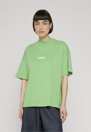 Kvinde med kort mørkt hår iført en lys grøn oversized T-shirt og mørke løse bukser mod en ensfarvet grå baggrund.