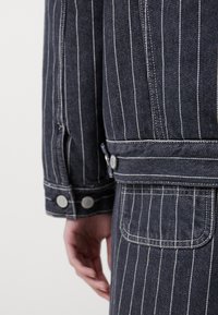 Spijkerjasje met een donkergrijze basis en witte strepen. Voorzien van zilveren knopen, een zak en zichtbare stiksels op de manchetten en de zoom.