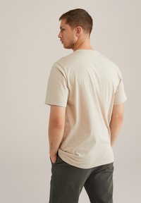 Beige, rövid ujjú póló, kerek nyakkivágással, puha tapintással és laza fazonnal, sötétzöld nadrággal párosítva. Látható tetoválás a bal alkarján.