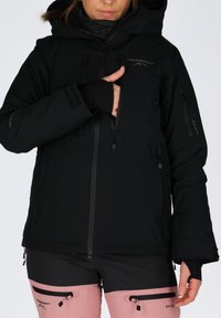 Black waterproof jacket with a hood, zippers, and multiple pockets; paired with pink pants featuring black accents and zippers.