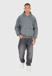 Homme portant un sweat à capuche gris délavé avec poche avant, un jean gris foncé ample et des baskets blanches devant un fond uni.