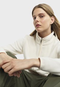 Veste zippée en polaire crème avec col montant, dotée d'une texture lisse et de manches longues, associée à un pantalon ample en olive.
