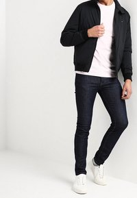 Giacca bomber nera sopra una maglietta rosa chiaro, abbinata a jeans aderenti blu scuro e sneakers bianche. Tessuto morbido e stile casual.