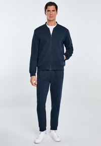 Giacca a zip in pile blu navy con scollatura rotonda, abbinata a pantaloni tapered coordinati. Presenta polsini a coste e sneakers bianche eleganti.