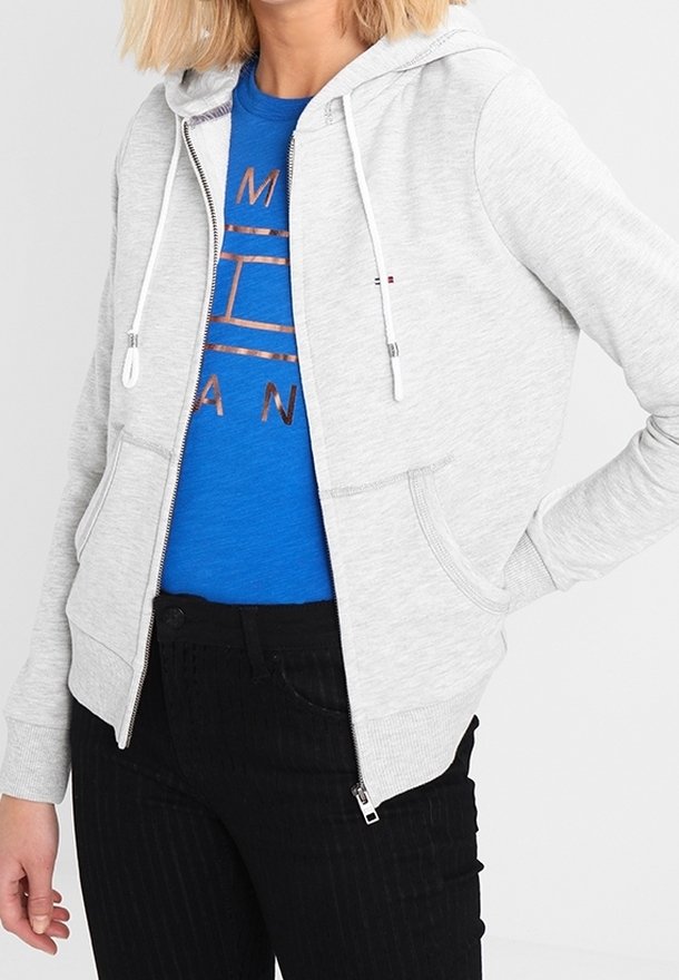 Sudadera con cremallera en un color gris claro, con capucha ajustable y bolsillos laterales. Se lleva sobre una camiseta azul con un diseño impreso, combinada con pantalones negros.