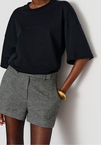 Černé oversized bavlněné tričko s krátkými rukávy, s jemným embosovaným textem. V kombinaci se šedými strukturovanými šortkami a zlatým náramkem.
