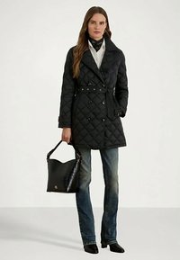 Mulher vestindo um casaco preto acolchoado com cinto, camisa branca, jeans azuis, botas pretas de tornozelo e segurando uma bolsa de couro preta.