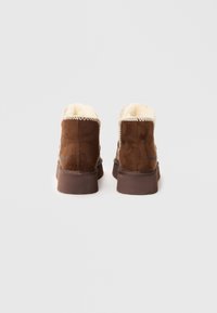 Bottines en daim marron avec doublure crème, semelle en caoutchouc et couture decorative le long du bord supérieur. Présente un design minimaliste.