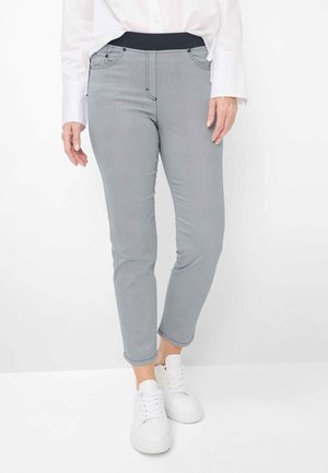 Kvinde iført tætsiddende grå bukser med mønster og sort linning, hvid skjorte med knapper og hvide snørebåndssko, stående.