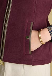 Burgunderfarbige Fleecejacke mit goldfarbenem Reißverschluss, kontrastierender grüner Borte und einer Seitentasche mit einem runden metallischen Akzent.
