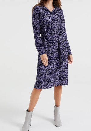 Mujer con vestido camisero abotonado con estampado de leopardo en púrpura y negro, con cinturón en la cintura y botas grises hasta el tobillo, de pie sobre un fondo blanco.