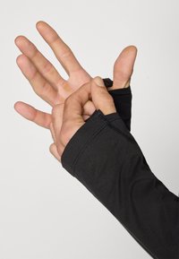 Svart tettsittende langermet genser med tommelhull, glatt tekstur, flate sømmer, som viser en håndgest og avdekker lagdelte fingre.