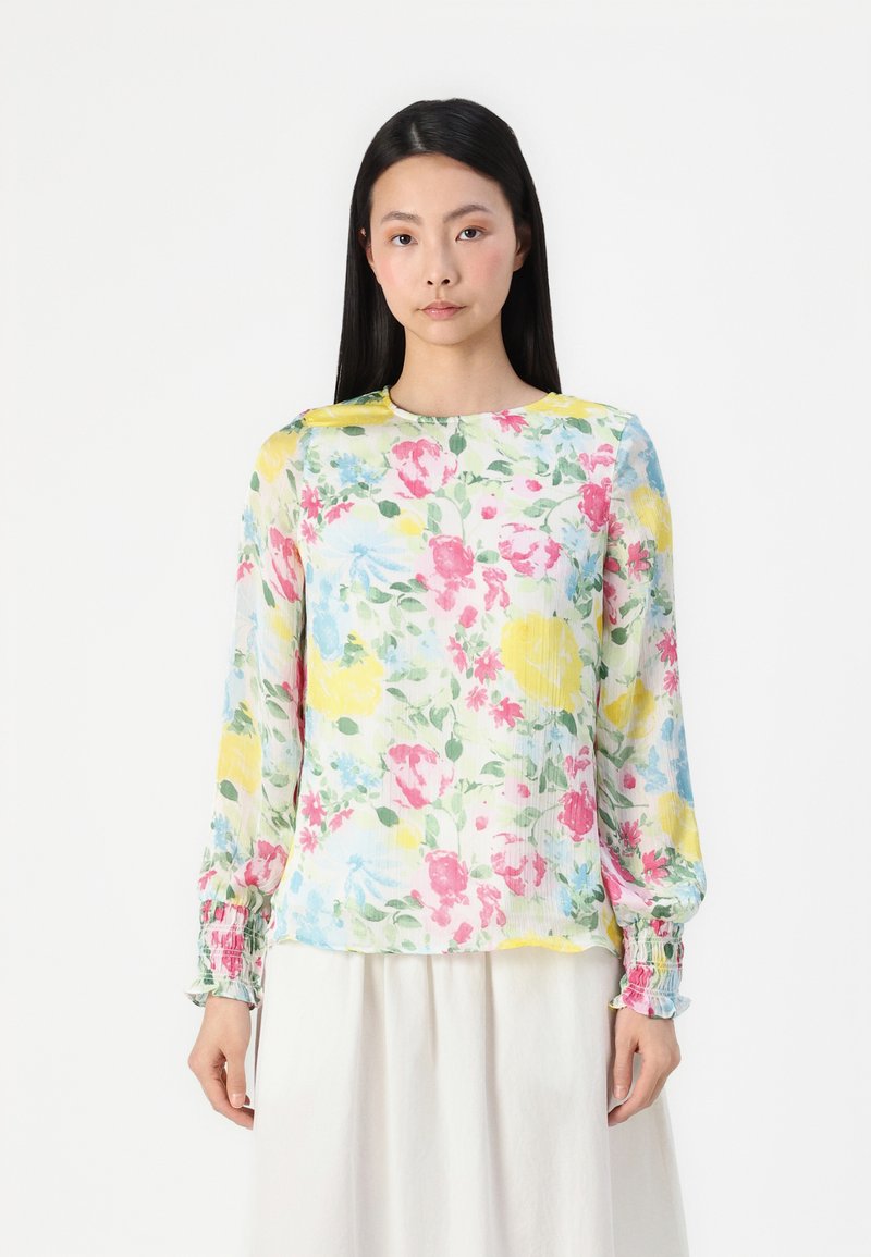 Blouse florale en tissu léger et transparent, mettant en avant un motif de fleurs roses, jaunes et bleues. Manches longues avec des poignets froncés.