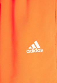 Chaqueta deportiva de color naranja brillante hecha de una tela suave, con un logo blanco de Adidas y un diseño de tres rayas en el lado izquierdo.