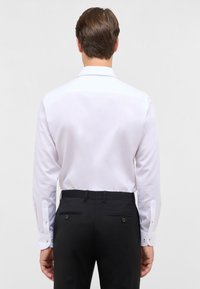 Camicia bianca a maniche lunghe in tessuto morbido con colletto classico, abbinata a pantaloni neri con due tasche posteriori e chiusura con bottone.