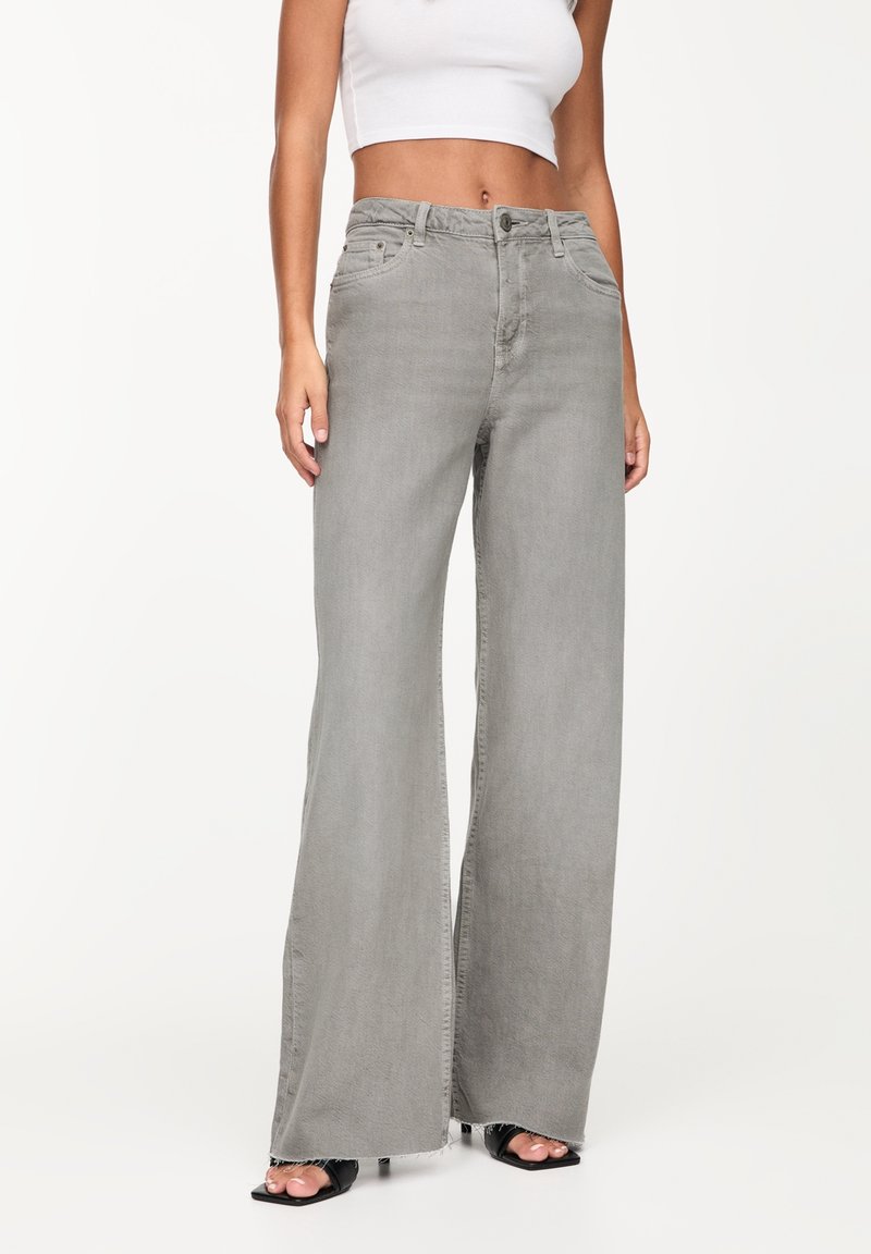 Femme portant un jean gris taille haute à jambes larges avec des ourlets bruts et des talons ouverts noirs, associé à un débardeur court blanc.