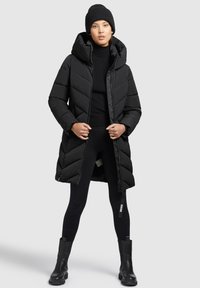 Zwarte pufferjas met capuchon, met chevronstiksels, ritsluiting en zijzakken, gedragen over een zwarte coltrui en legging.