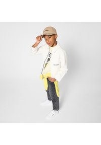 Λευκό τζιν μπουφάν, κίτρινο φούτερ δεμένο στη μέση, μπλουζάκι με γραφικά, γκρι τζιν, και λευκά αθλητικά παπούτσια· μπεζ καπέλο με λεπτομέρεια λογότυπου.