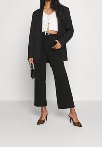 Schwarzer Blazer über einem weißen Cropped-Pullover mit Bindeband, hoch taillierte schwarze weitgeschnittene Jeans und High-Heel-Mules im Leopardenmuster. Kleine schwarze Handtasche.