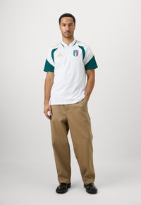 Hvit polo skjorte med grønne ermer, glidelås-kollared, Italia-emblem og gullakenter. Kombinert med khaki-bukser og sorte sko.